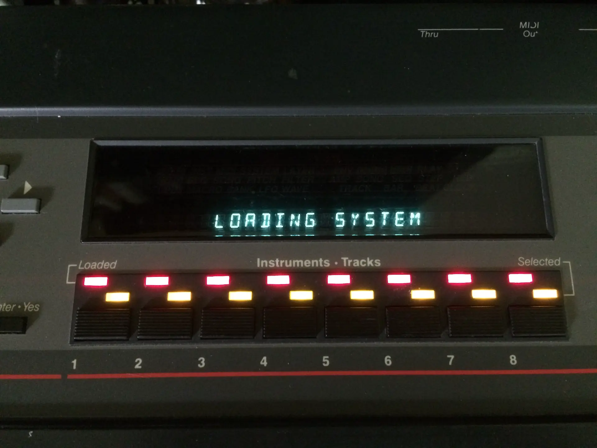 Late-1980s black digital sampler unit with small green LCD display, grey buttons and a data wheel, photographed against a grey archival background