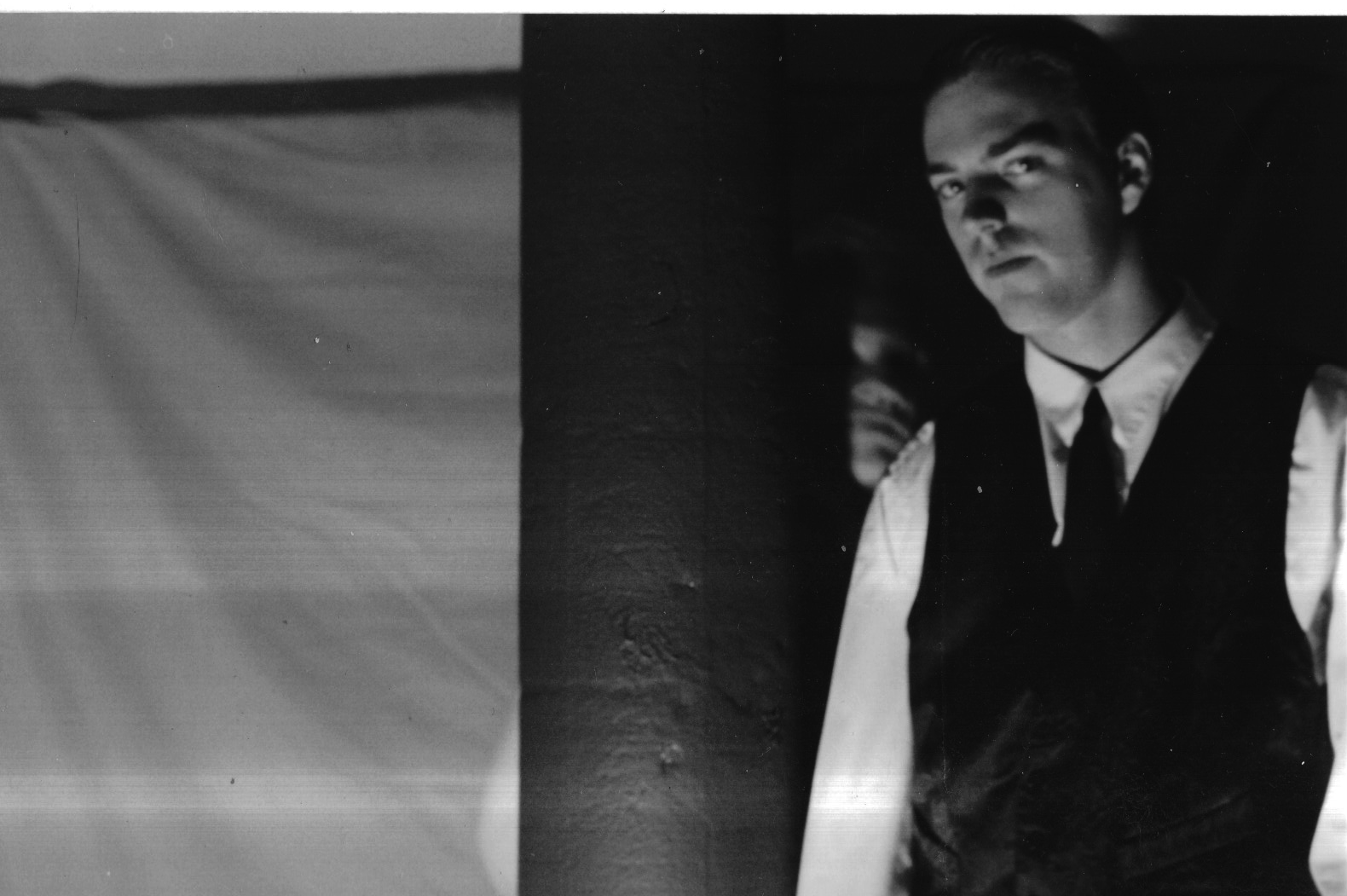 Distressed black-and-white photograph of Sidney in a dark vest and tie standing beside a partition, half his face lit, light banding across the frame